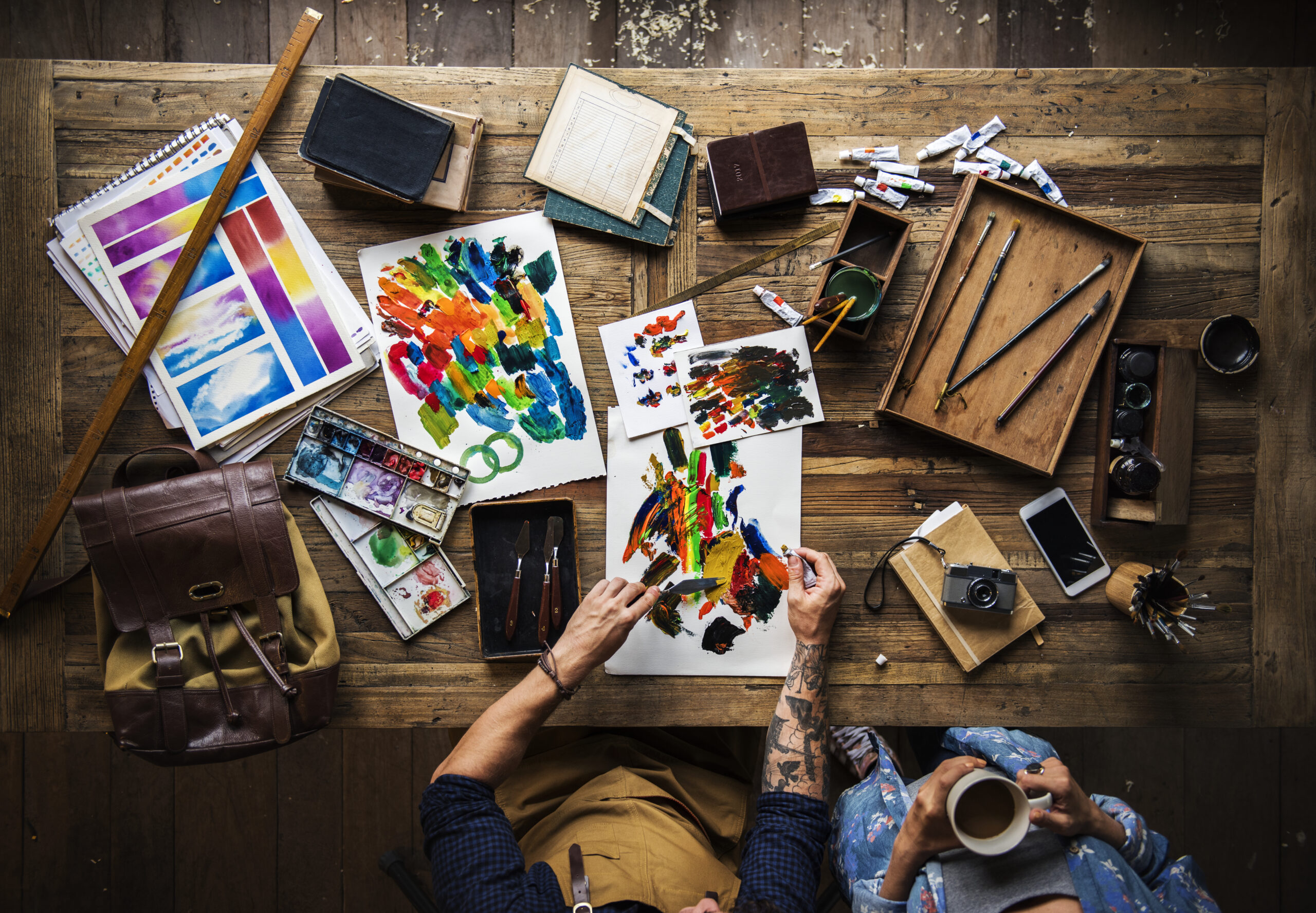 Dos personas pintando sobre una mesa llena de materiales artísticos como pinceles, pinturas acrílicas, cuadernos, espátulas y una cámara antigua.