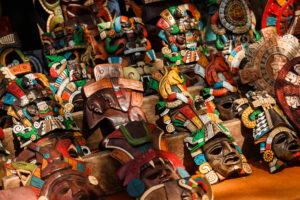Máscaras de madera pintadas a mano con motivos mayas y aztecas, dispuestas sobre estantes de madera en un mercado local.