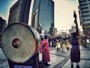 Grupo de personas en trajes tradicionales coreanos participando en una ceremonia frente a modernos rascacielos en una ciudad, con un gran tambor decorativo en primer plano.