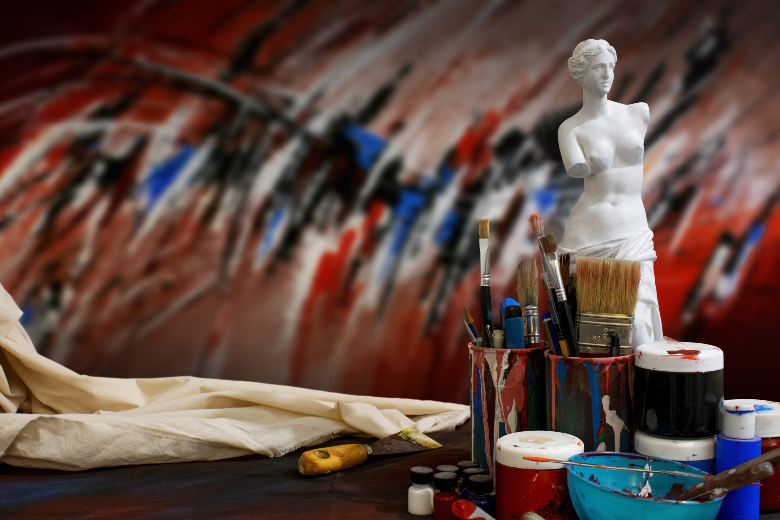Pinceles y frascos de pintura frente a una escultura de Venus de Milo, con fondo abstracto en tonos rojos, negros y azules.