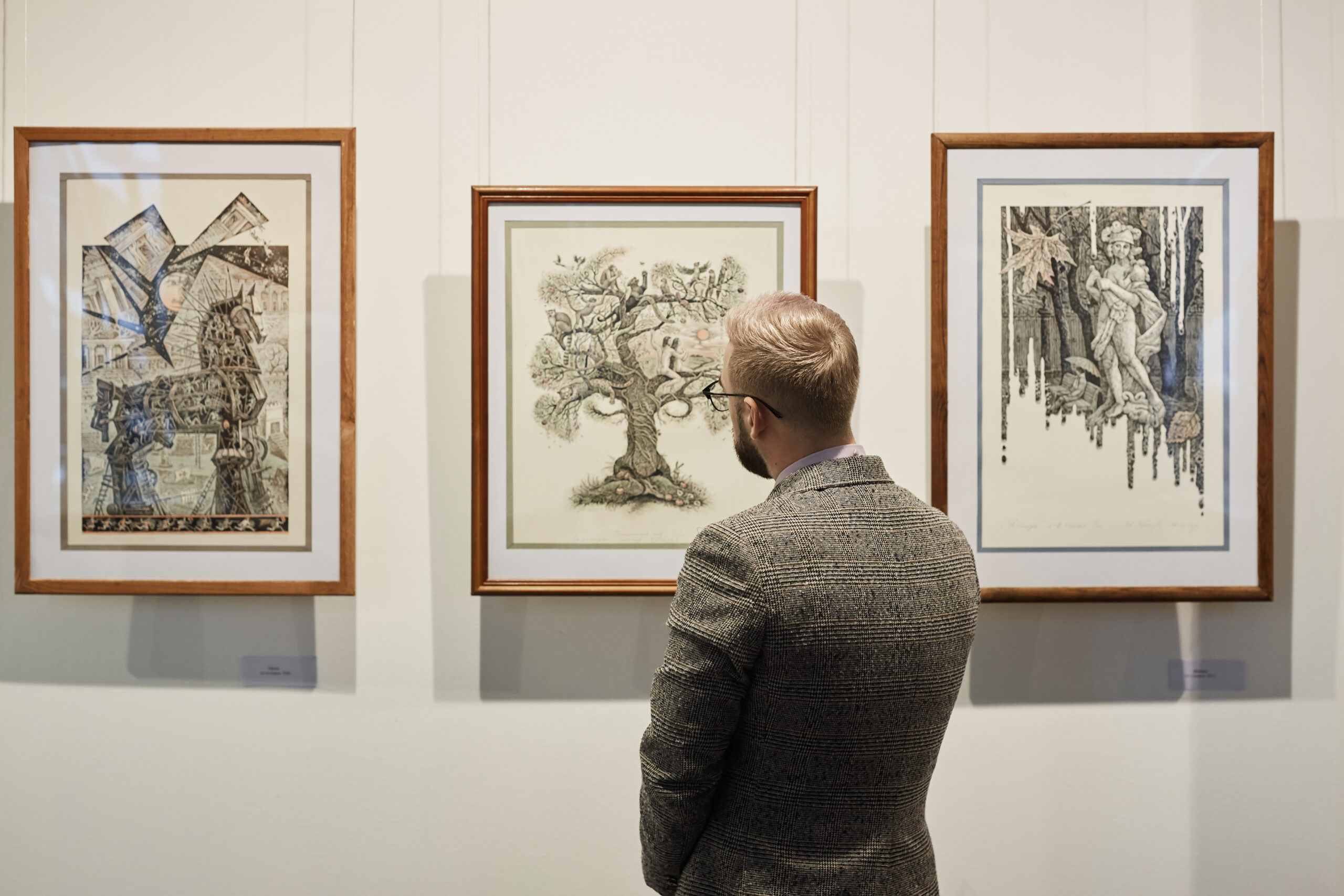 Hombre de espaldas observando tres obras de arte moderno enmarcadas colgadas en una pared de galería.
