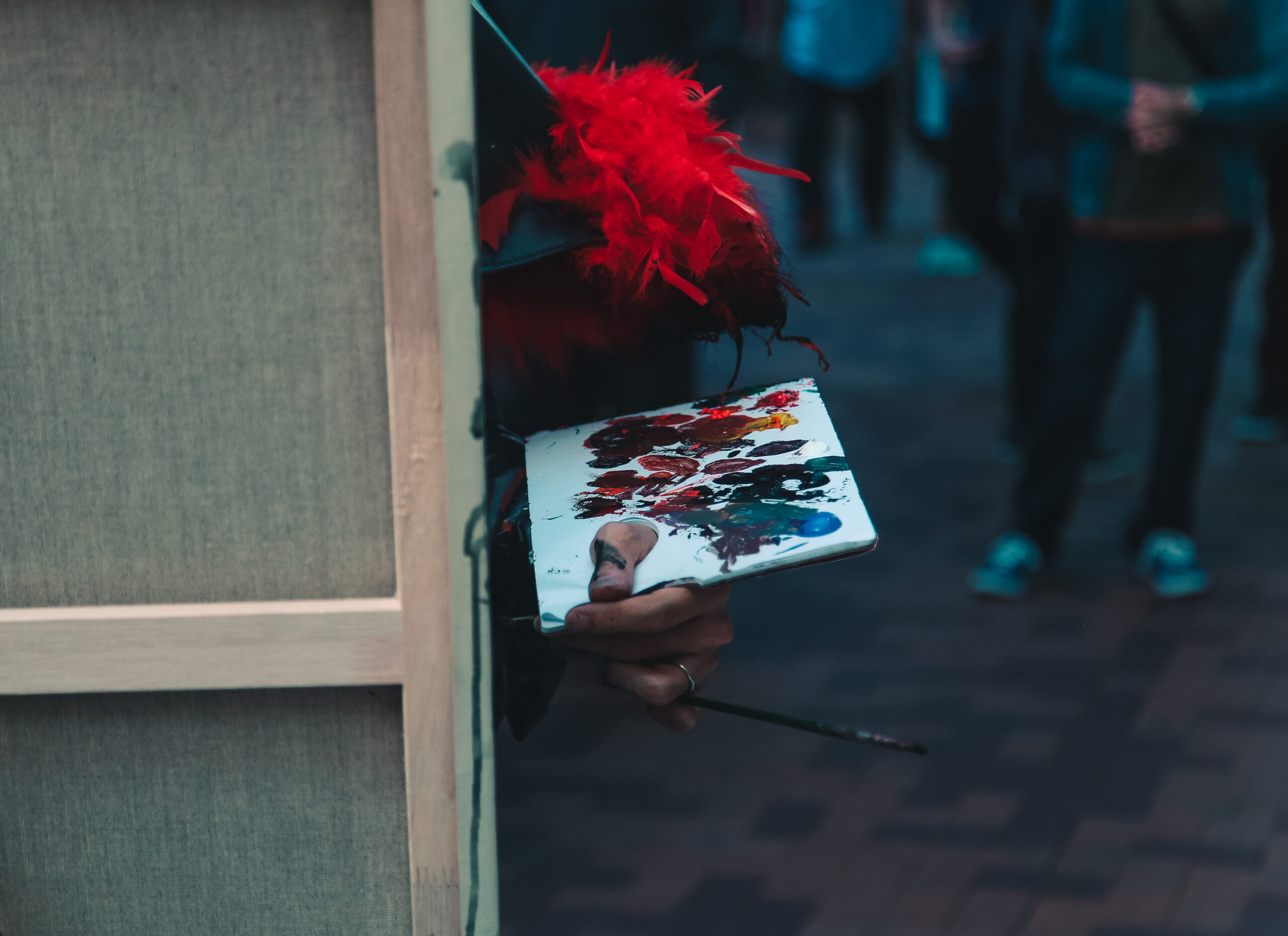 Mano de un pintor callejero sosteniendo una paleta con múltiples colores y un pincel, detrás de un lienzo, con una pluma roja decorativa en el brazo.