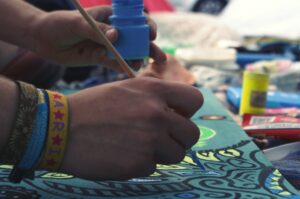 Mano pintando con pincel azul sobre una tela con diseño artístico, rodeada de materiales de arte y pulseras coloridas.