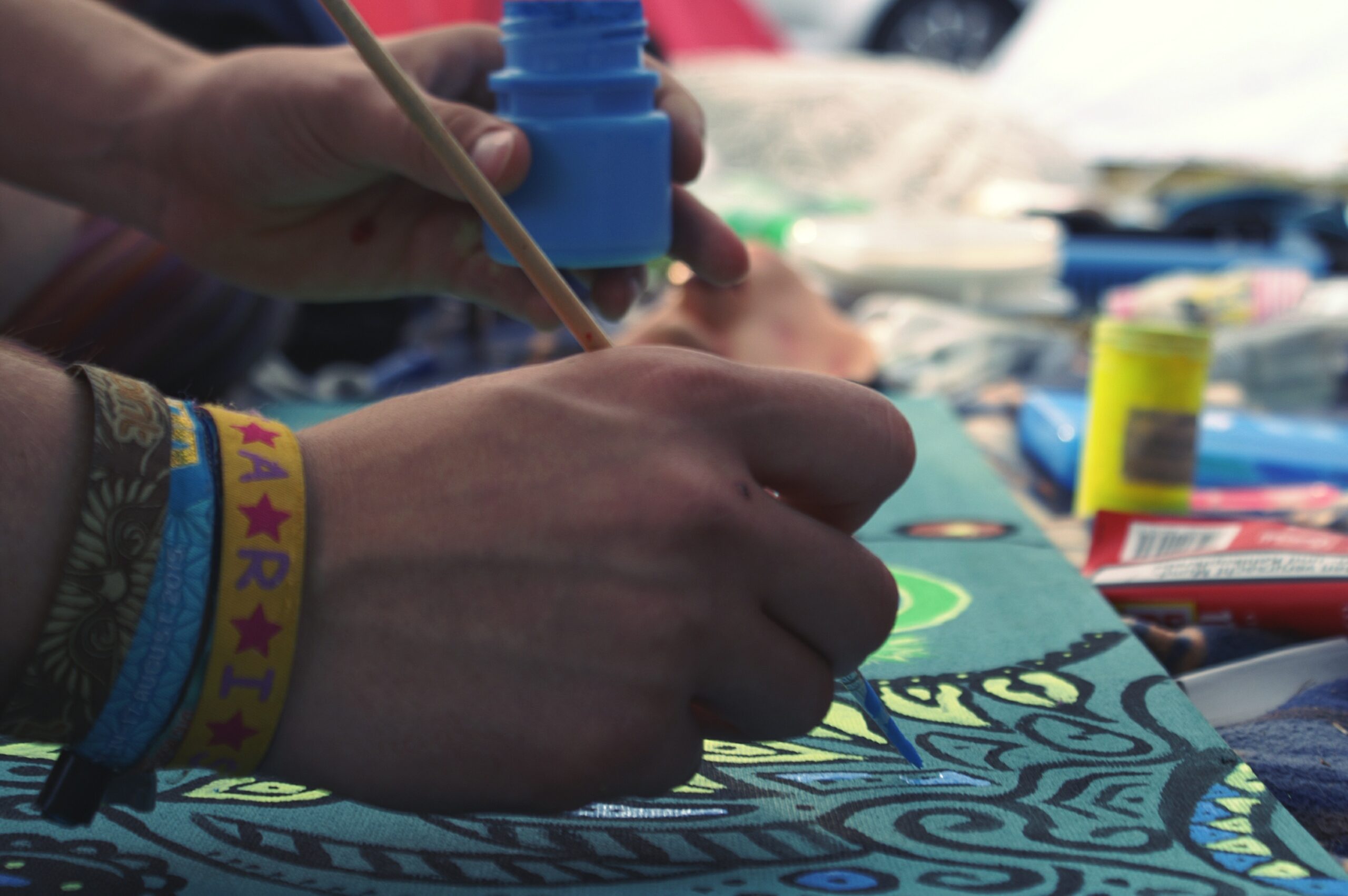 Mano pintando con pincel azul sobre una tela con diseño artístico, rodeada de materiales de arte y pulseras coloridas.