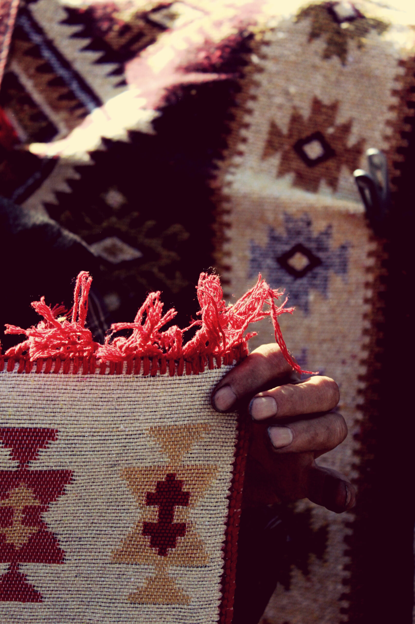Mano de una persona sosteniendo un tejido artesanal con flecos rojos y patrones geométricos, sobre un fondo de telas similares.