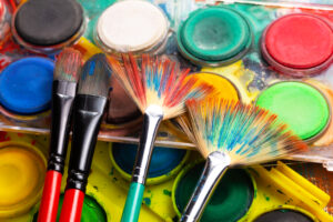Varios pinceles, incluidos abanico y planos, reposan sobre una paleta de acuarelas con colores intensos como rojo, azul, verde y amarillo.