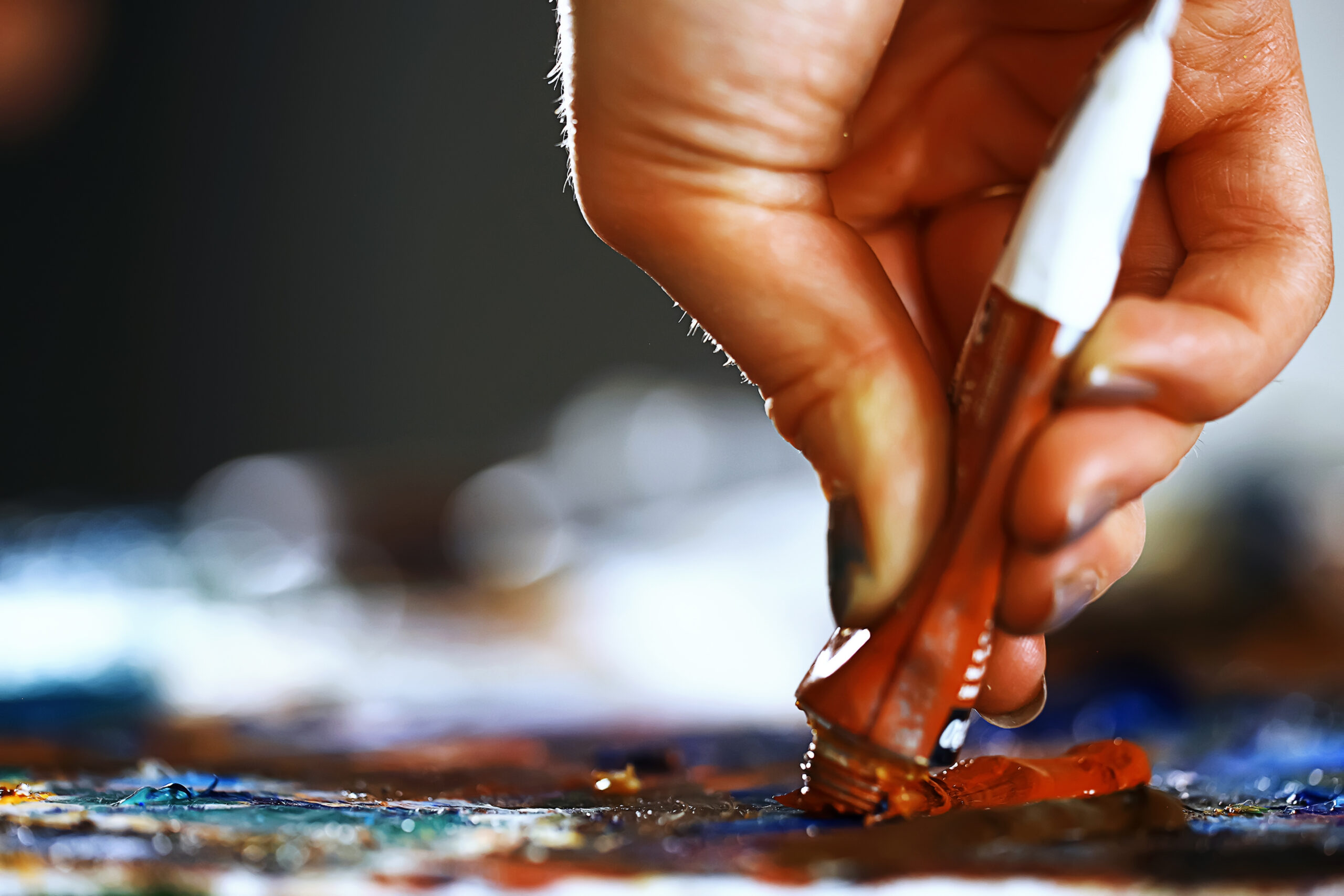 Mano presionando un tubo de pintura al óleo sobre una superficie llena de colores, con pigmento naranja saliendo y textura visible en primer plano.