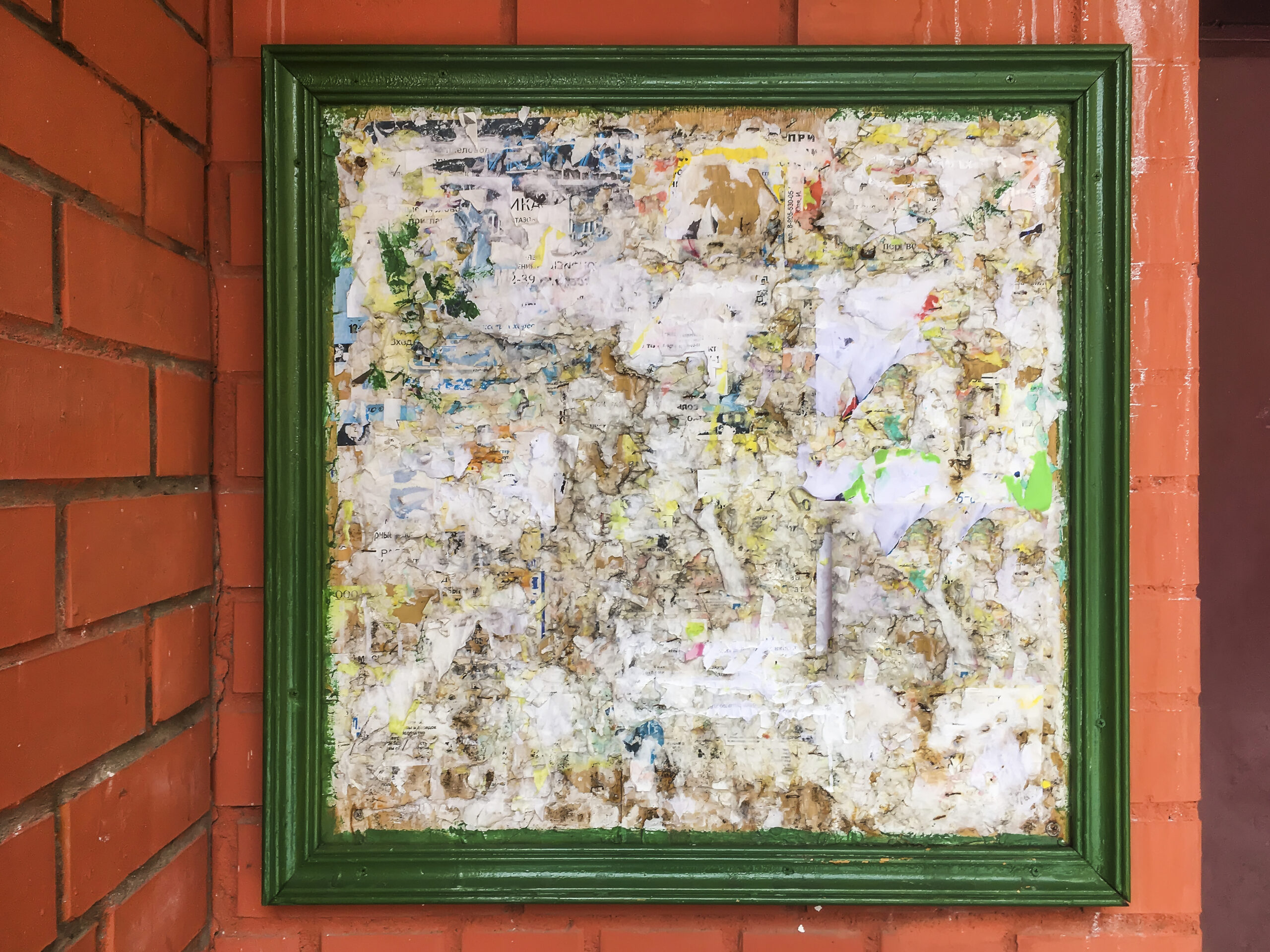 Tablón de anuncios con marco verde cubierto de restos de papel y carteles desgastados, colocado sobre una pared de ladrillos rojos.