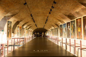 Corredor interior de la Venaria Reale en Turín, Italia, con techo abovedado de ladrillo e iluminación cenital, flanqueado por retratos de figuras históricas colgados en ambos lados.