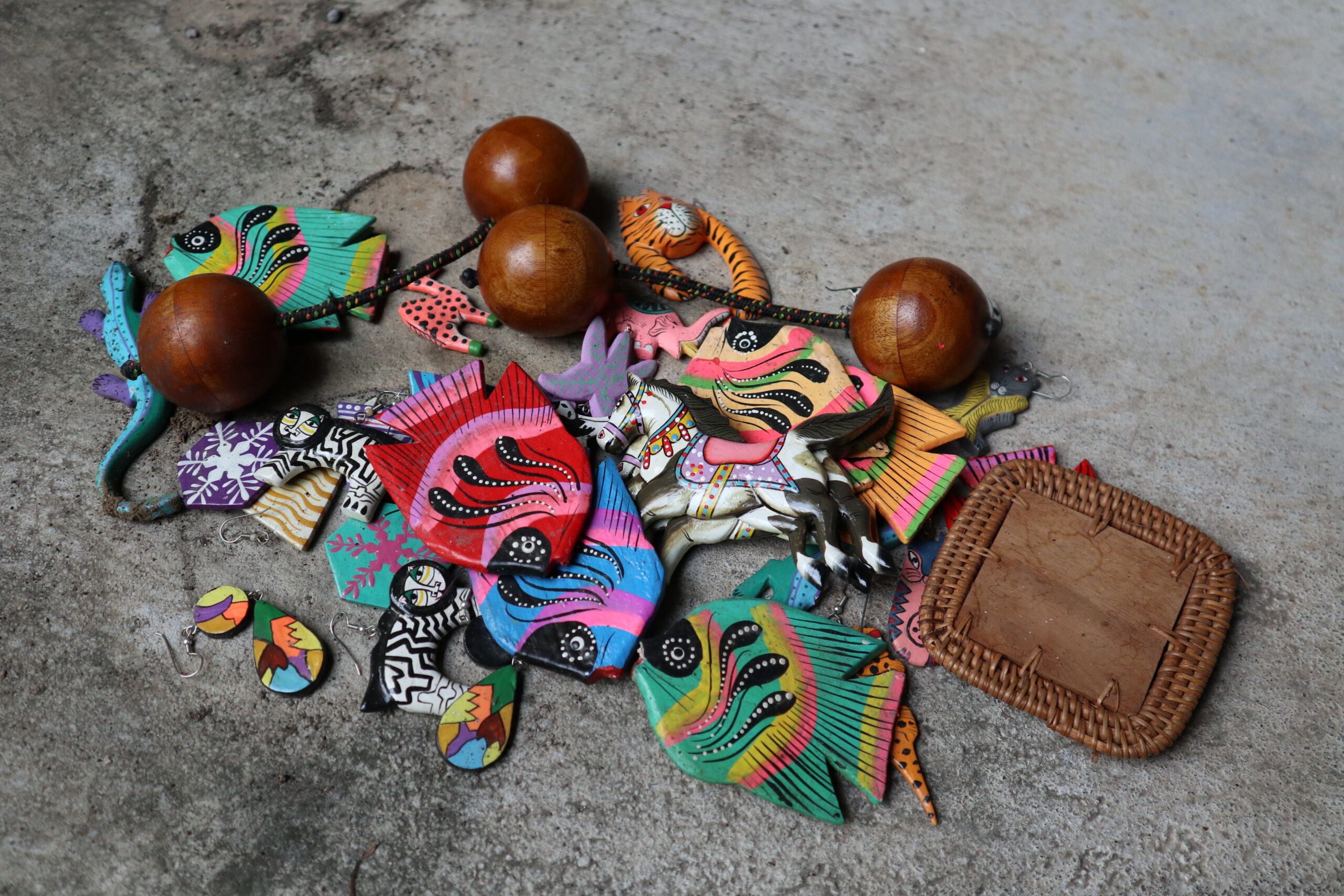 Artesanías de madera pintadas con colores brillantes, incluyendo peces, caballos y figuras de animales, junto a aretes y un marco de palma sobre una superficie de concreto.