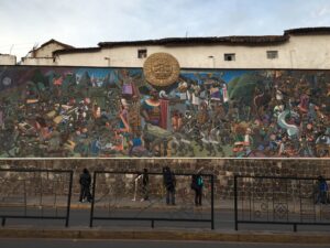 Mural histórico en una calle de Cusco que muestra una batalla entre incas y conquistadores españoles, con detalles coloridos y simbólicos de la cultura andina, bajo un medallón dorado en el centro superior.