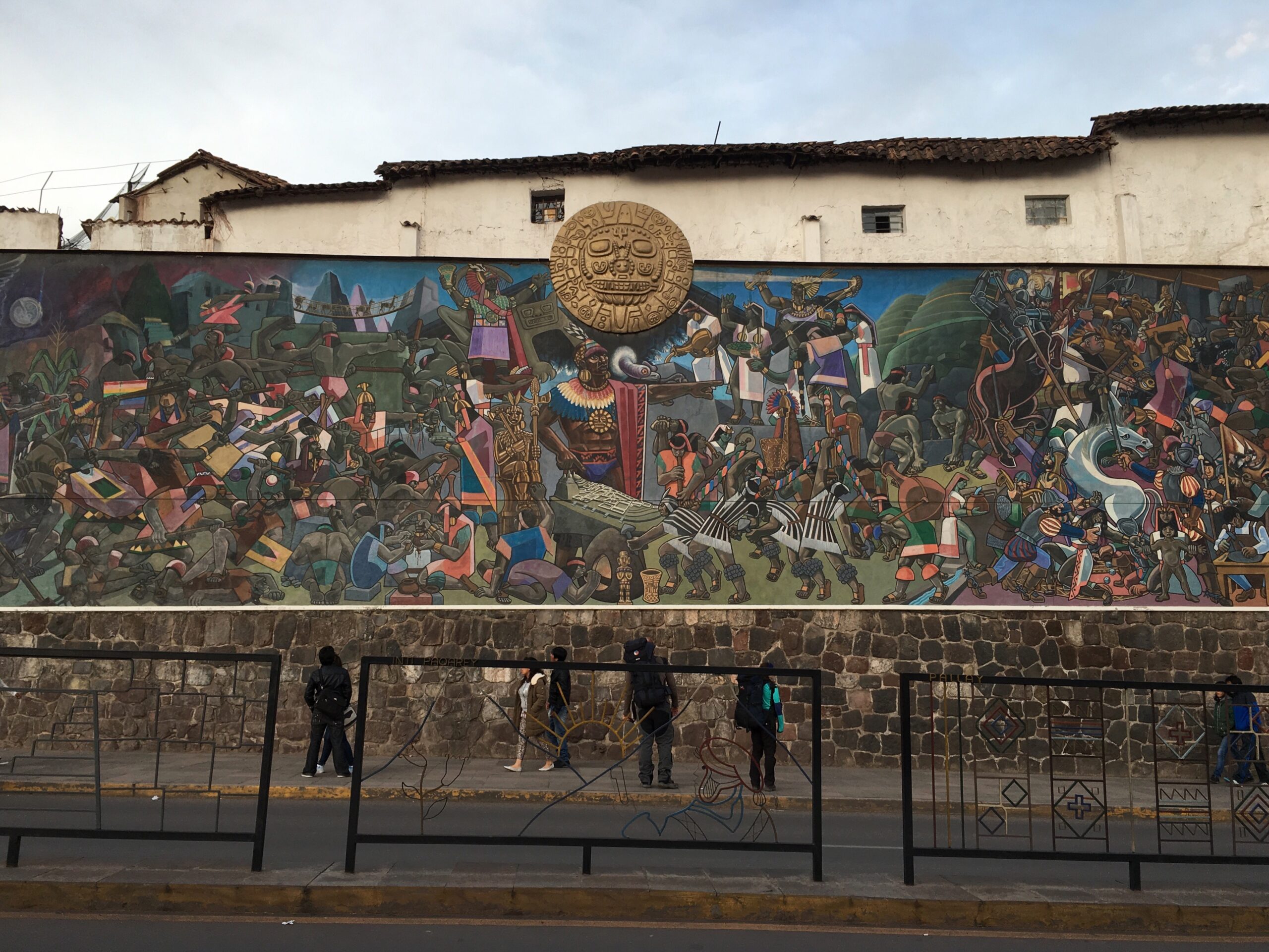 Mural histórico en una calle de Cusco que muestra una batalla entre incas y conquistadores españoles, con detalles coloridos y simbólicos de la cultura andina, bajo un medallón dorado en el centro superior.
