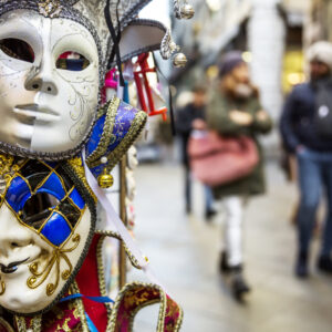 Máscaras coloridas y decoradas del Carnaval de Venecia colgadas en un puesto, con personas caminando desenfocadas al fondo.