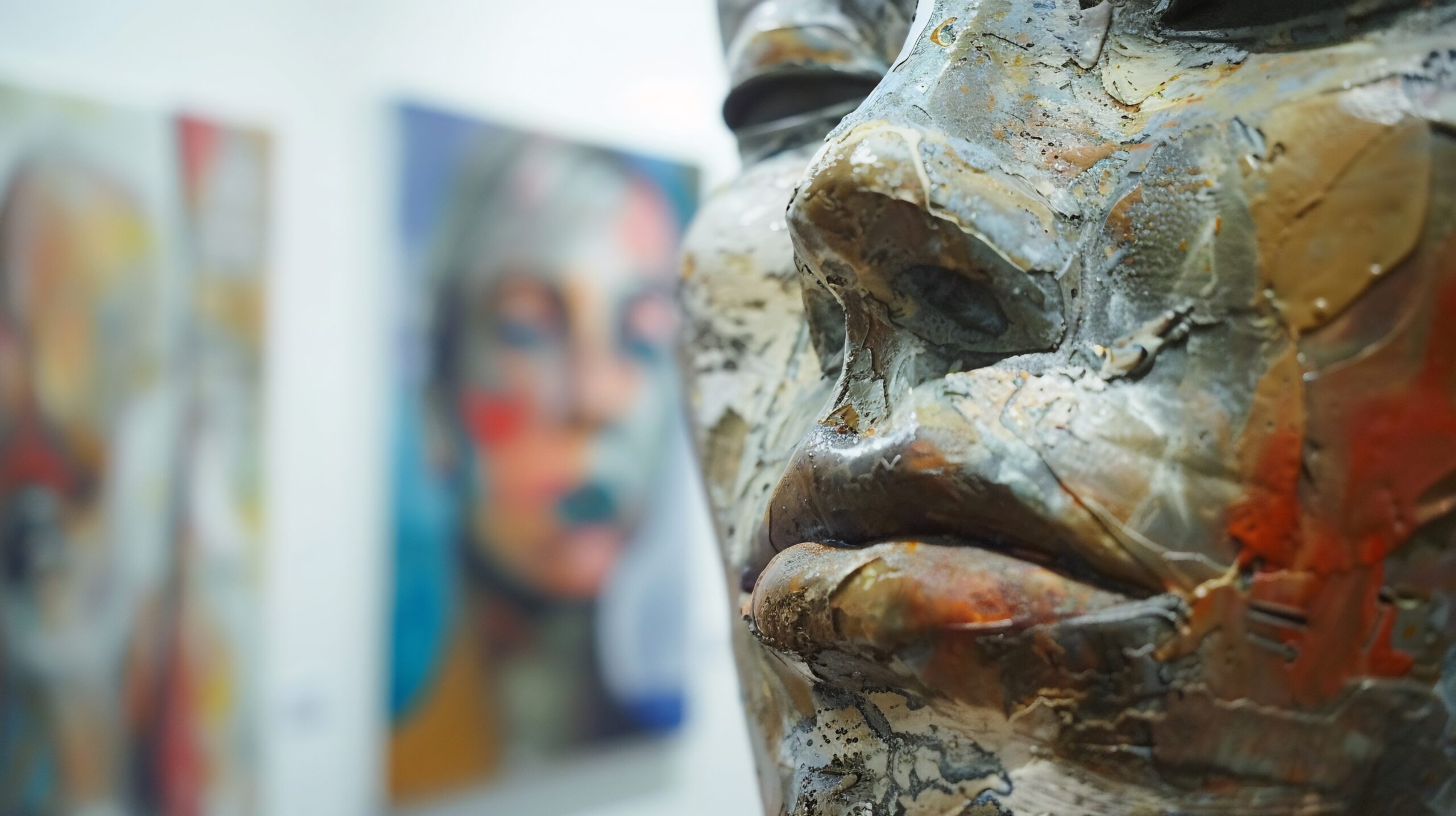 Primer plano de una escultura de rostro humano con texturas rugosas y colores terrosos, exhibida en una galería con pinturas abstractas desenfocadas al fondo