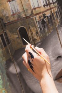 Mano con manchas de pintura sosteniendo un pincel frente a un mural que muestra una fachada antigua en tonos cálidos.